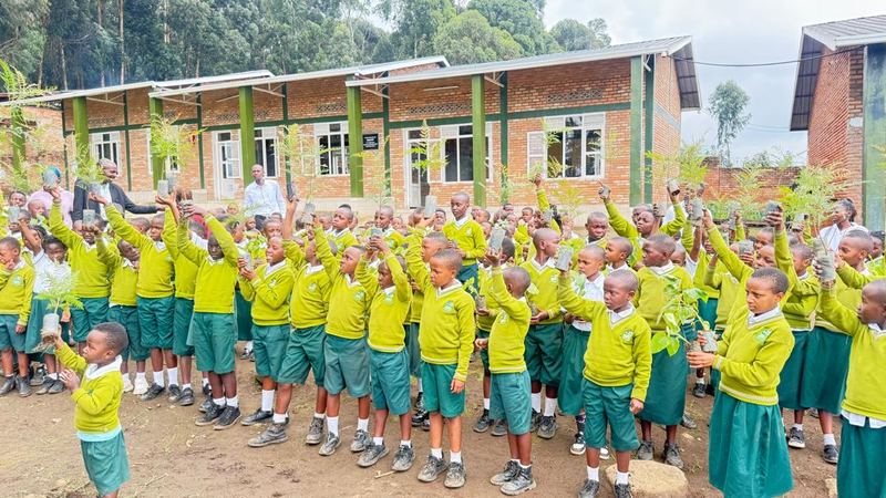 A large crowd of pupils in yellow jumpers and green uniform raising their arms in the yard in front of the brick classroom blocks