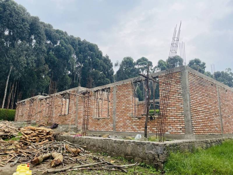 A brick classroom block under construction, with the roof frame up and tall trees behind