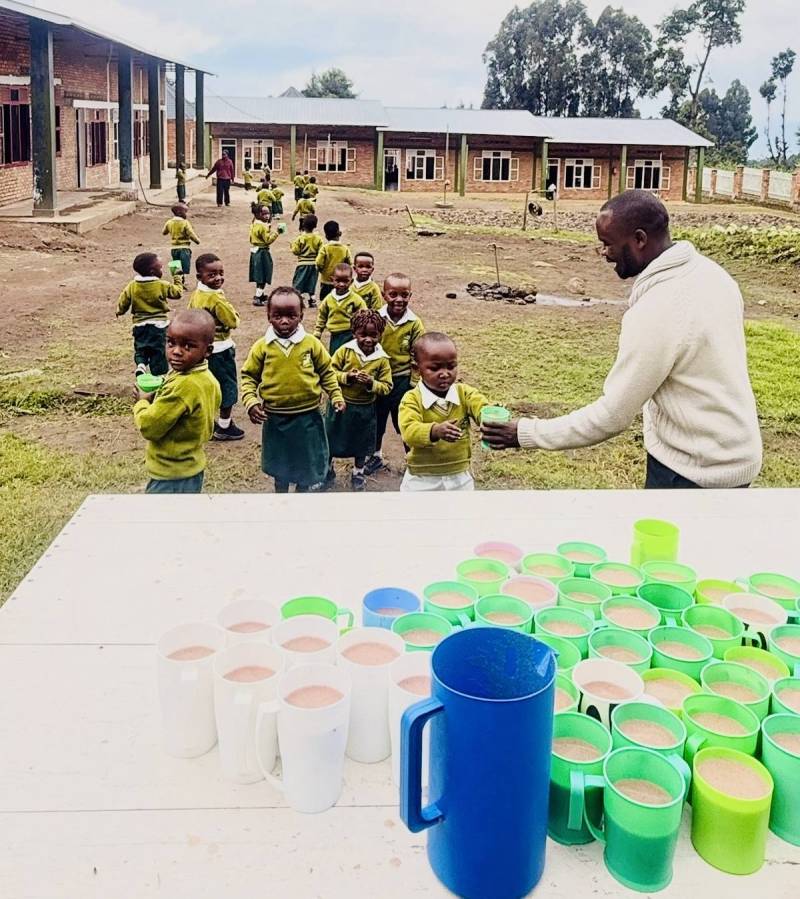 A staff member handing a cup to a young pupil as children in green uniform queue in the school yard, with rows of filled cups on a table