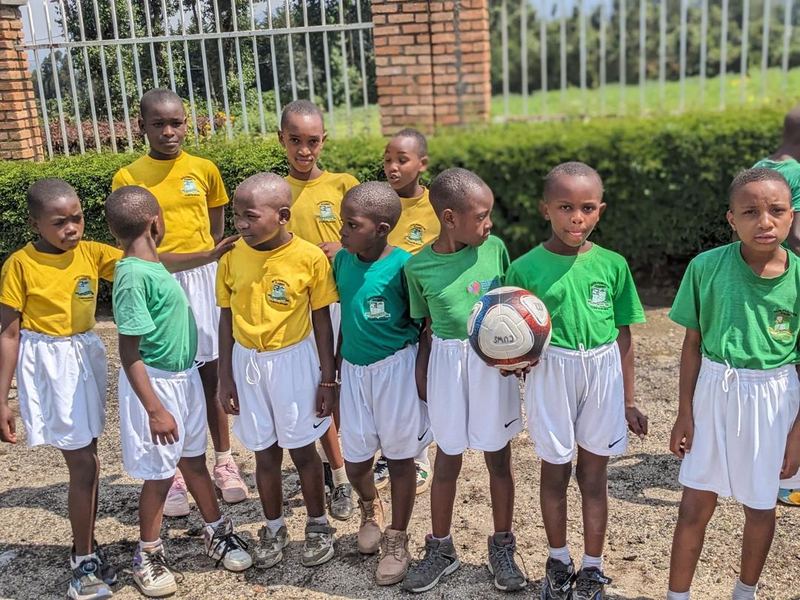 Pupils in yellow and green shirts and white shorts standing on a dirt yard, two of them holding footballs