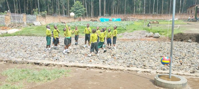 Pupils in yellow and green uniform playing on a gravel yard beside a flagpole, with forest behind