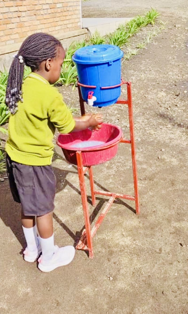A young pupil in a green school jumper washing her hands at a red basin under a blue water tank with a tap