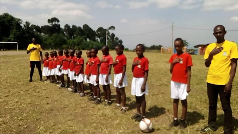 Two lines of pupils, one in red shirts and one in yellow, standing with hands on hearts on a grass field, a football at their feet