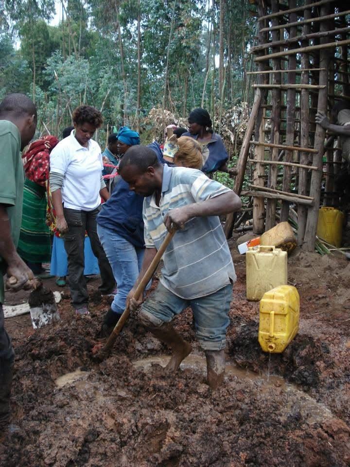 A man digging wet mud with a hoe as women look on, yellow jerrycans beside them