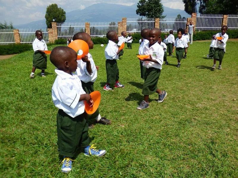 Young pupils in white shirts and green shorts playing with orange cones on a grass lawn, mountains behind