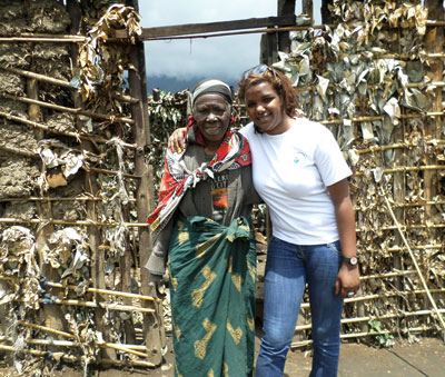 Faith Uwantege in a white T-shirt with her arm around an older woman in a headscarf and green wrap, in front of a wall of woven branches