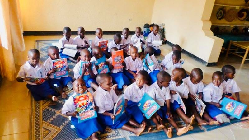 Young pupils in white shirts and blue shorts sitting on a patterned mat in a room, each holding up a picture book