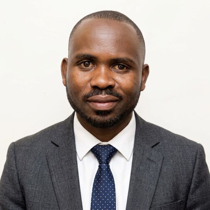 Asiimwe Pathias in a grey suit, white shirt and navy spotted tie, against a white background