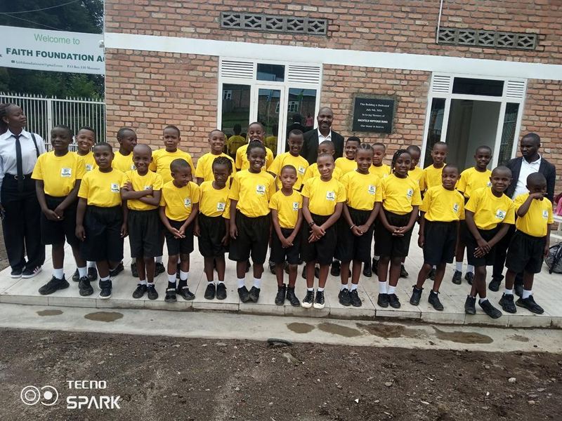 A row of pupils in yellow shirts and black shorts with staff, outside a brick building with a Faith Foundation sign