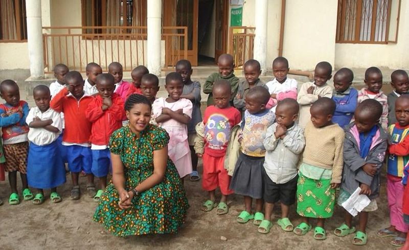 Faith Uwantege, the founder, crouching in a green patterned dress in front of a large group of young children with folded arms outside the school
